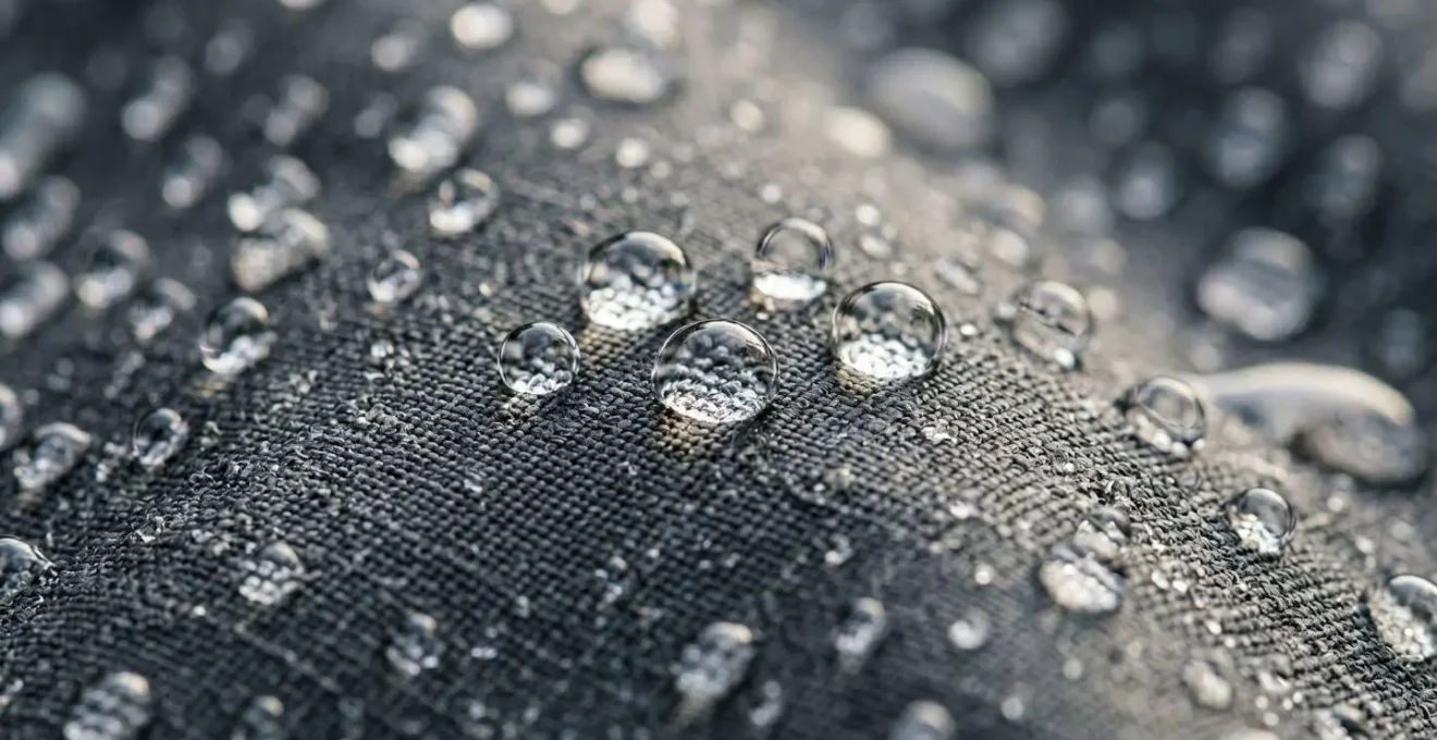 Dettaglio macro di tessuto tecnico impermeabile con gocce d'acqua che scivolano sulla superficie mostrando la texture e le proprietà idrorepellenti